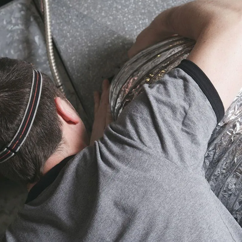 A person wearing a headlamp working on flexible metallic ducting in a crawl space.